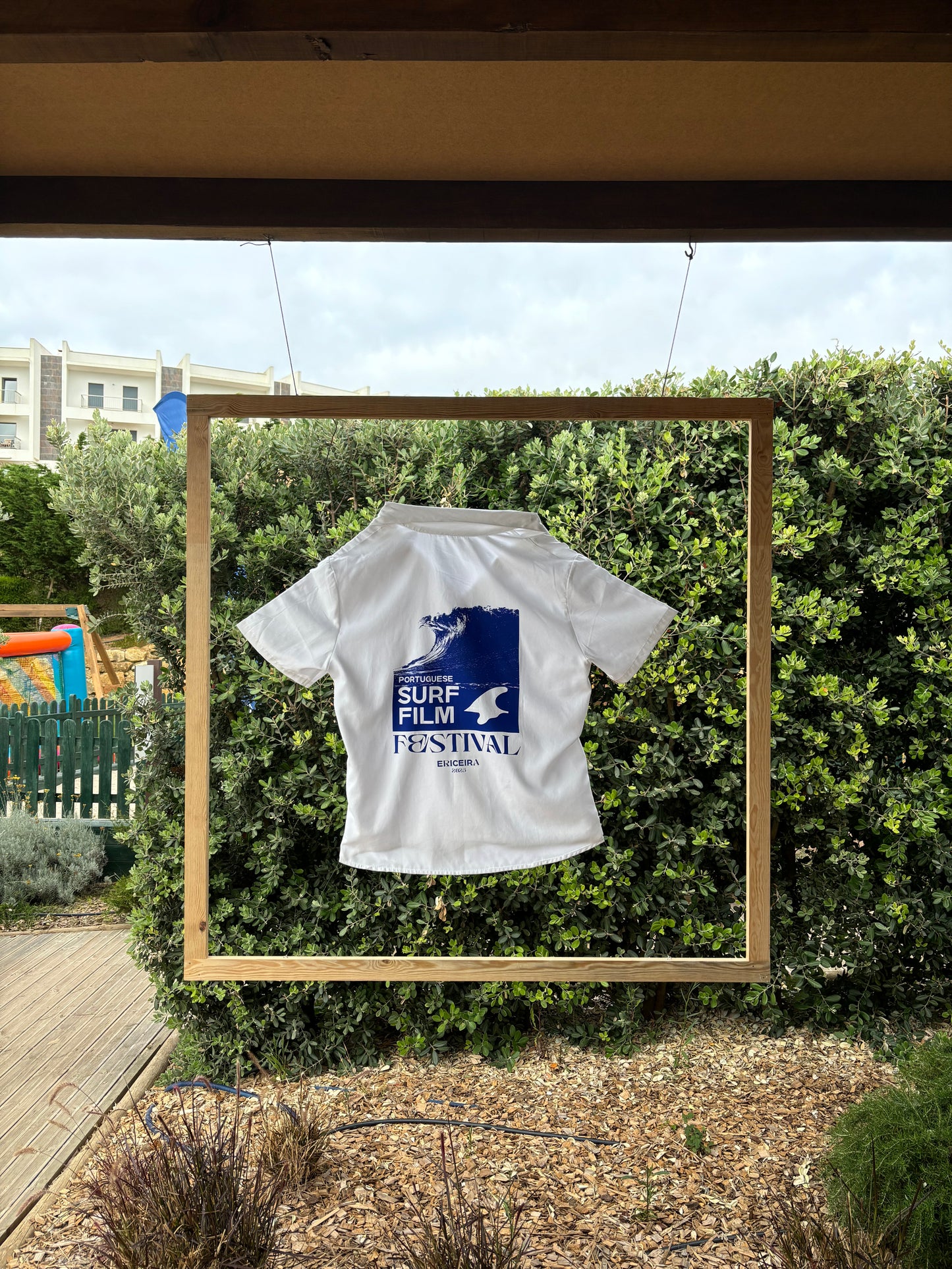 White shirt with blue graphic design on a wooden frame against a green bush background