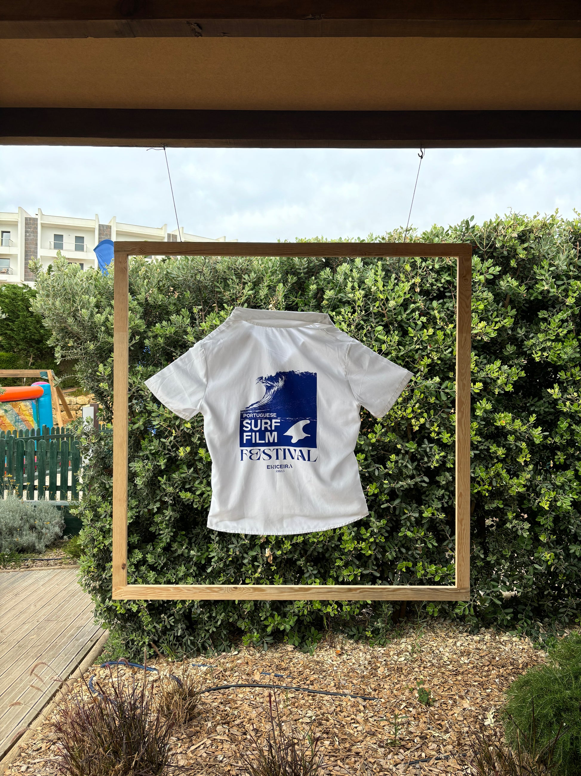 White shirt with blue graphic design on a wooden frame against a green bush background